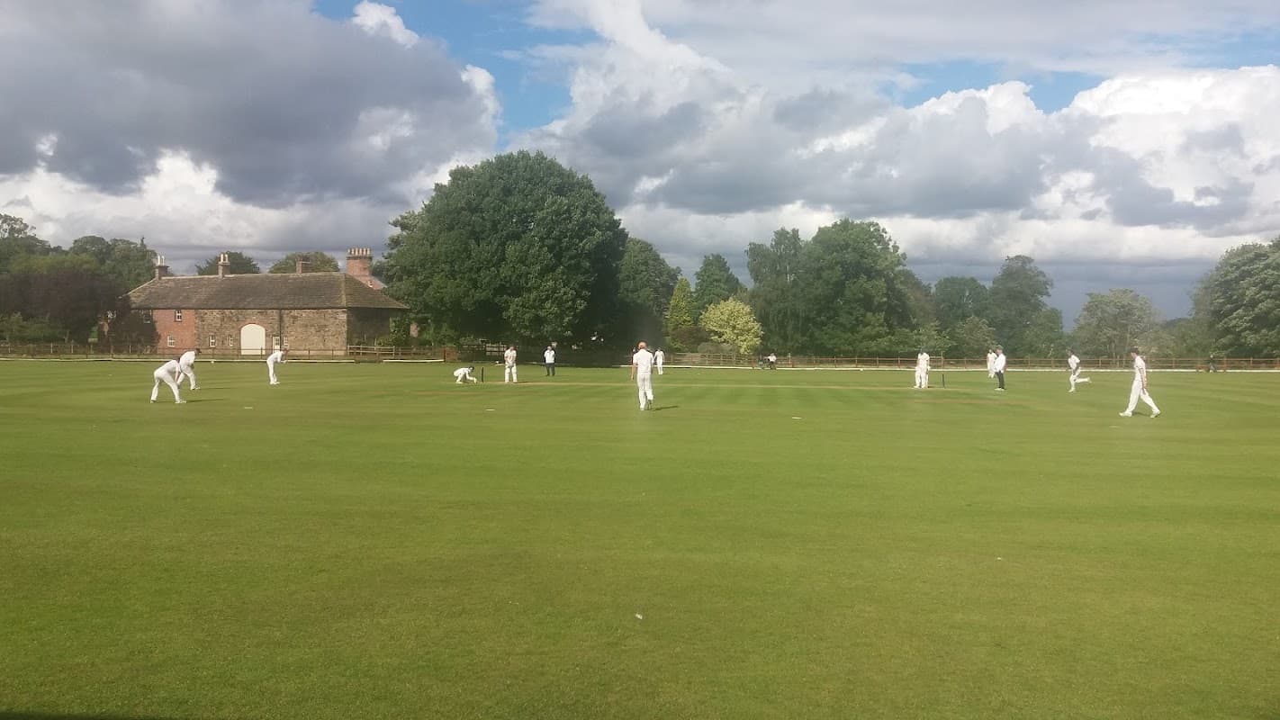 Cricket match in progress on a lush green field with players in white uniforms and a historic building in the background.