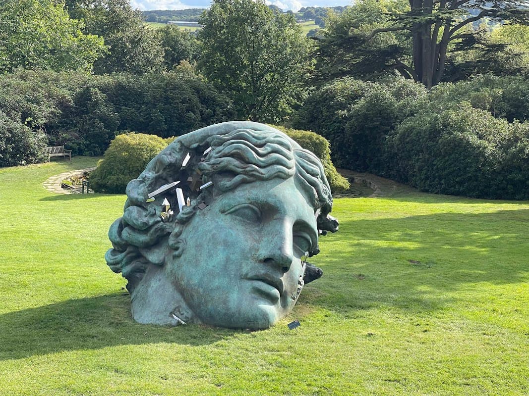 Large bronze sculpture of a head in a grassy area, surrounded by trees and shrubs at Yorkshire Sculpture Park.