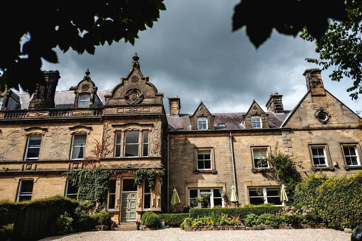 Victorian-style manor with ornate stonework, lush greenery, and outdoor seating under umbrellas against a cloudy sky.