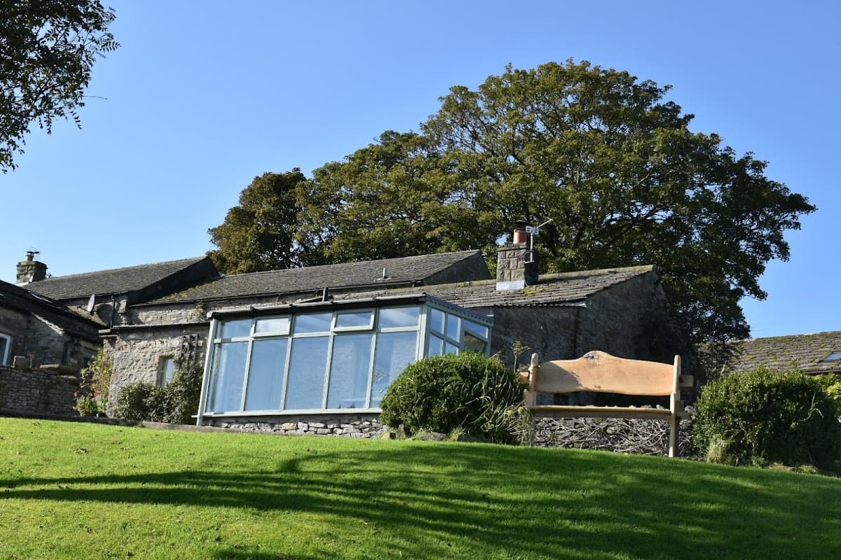 West Close Farmhouse features a stone exterior, large windows, and lush green surroundings under a clear blue sky.