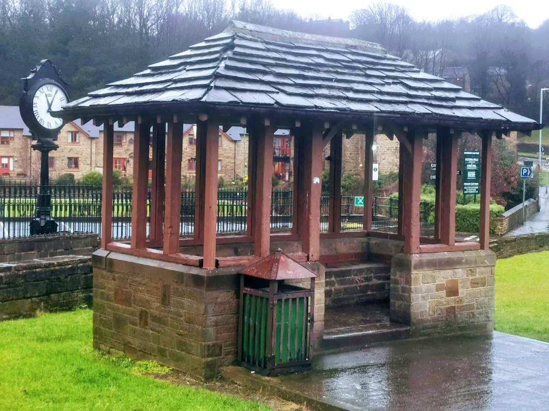A wooden gazebo with a slate roof, next to a clock and a green waste bin, in a grassy area of a car park.