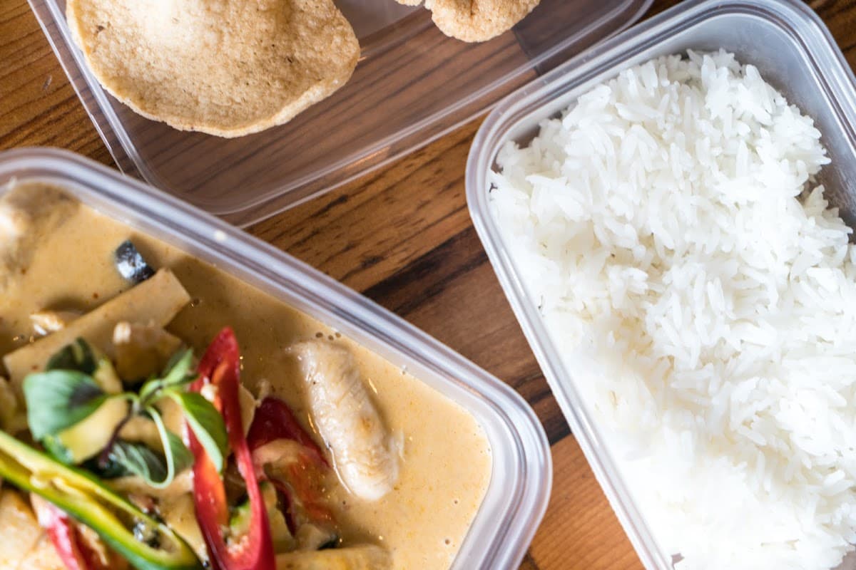 Thai curry with vegetables, served with white rice and crispy snacks in takeaway containers on a wooden surface.
