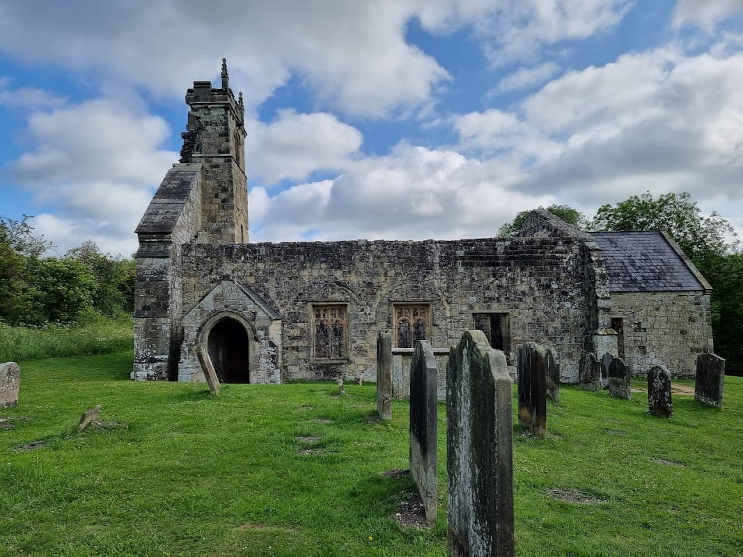 Ruined church with a tower, surrounded by gravestones and lush greenery under a cloudy sky.