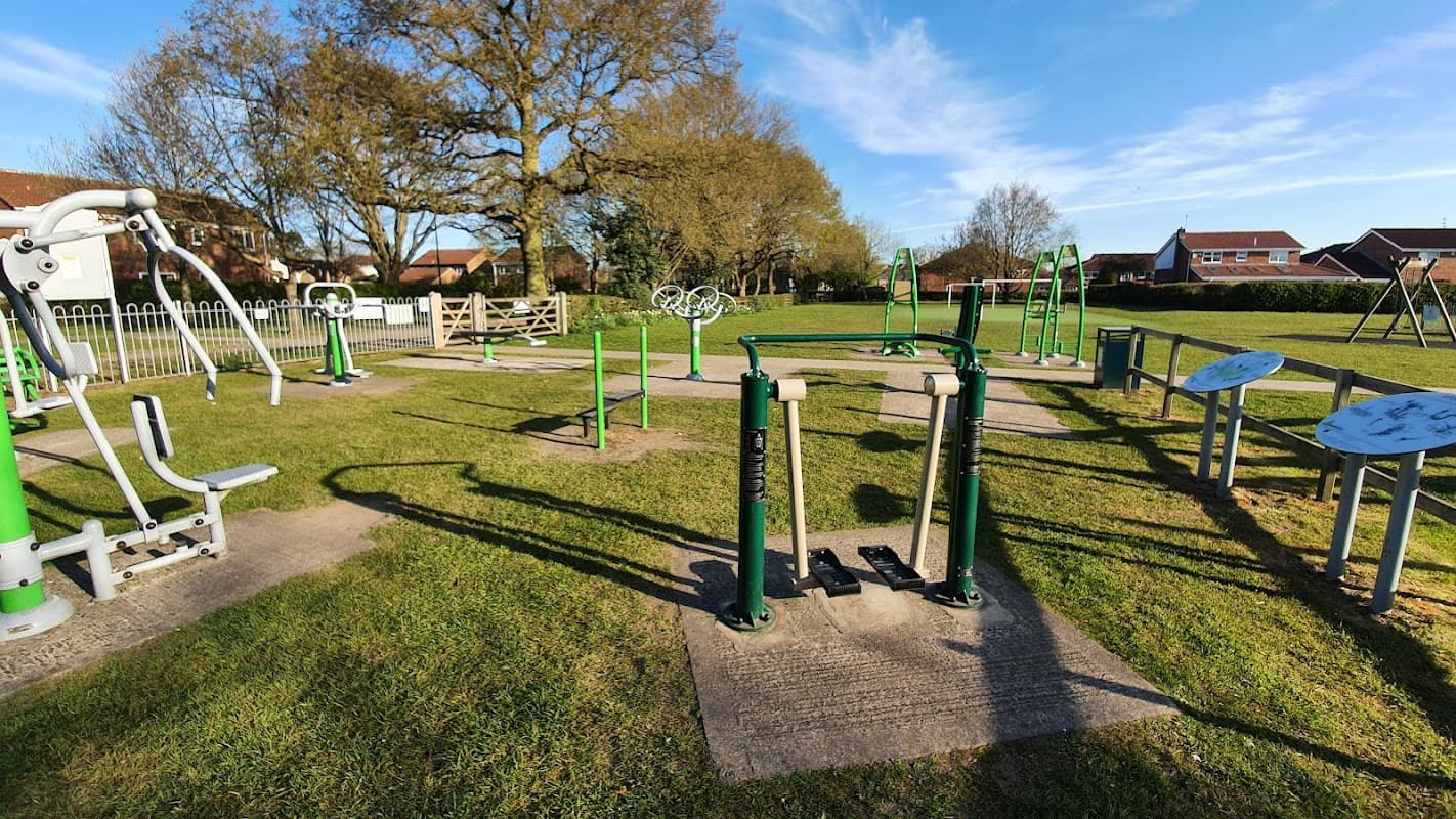 Outdoor fitness area with exercise equipment, grassy space, trees, and benches under a clear blue sky.