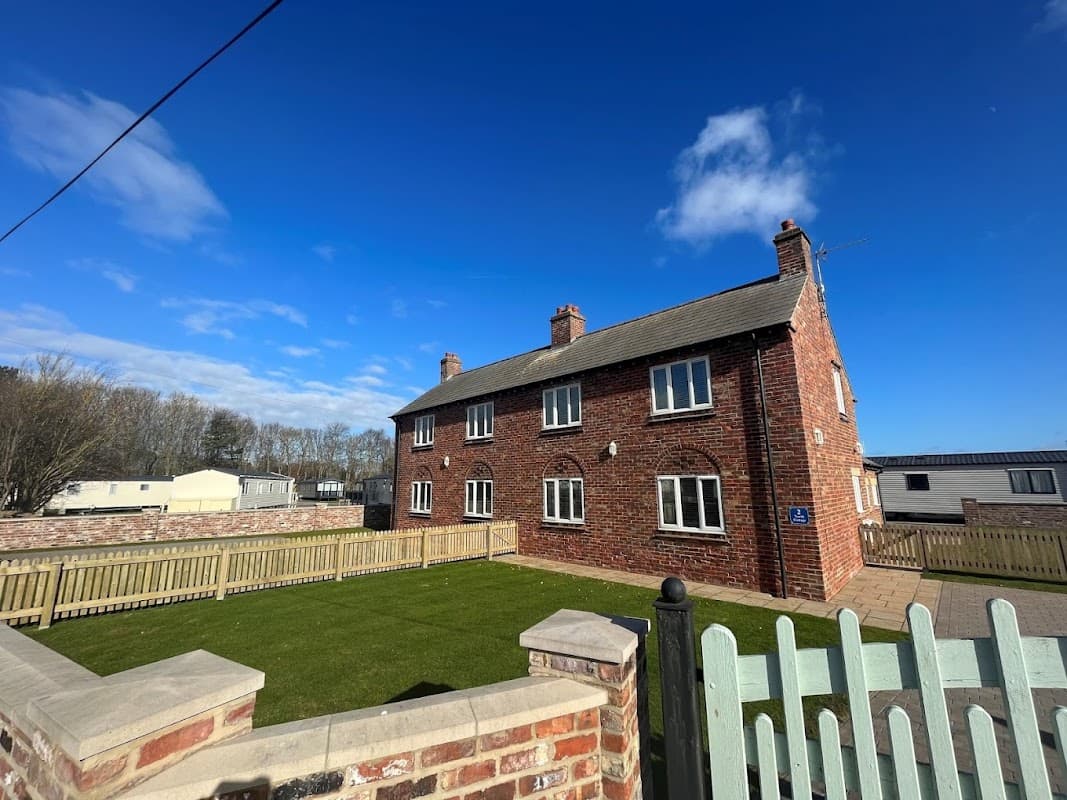 Red brick building with multiple windows, surrounded by a green lawn and wooden fence under a clear blue sky.