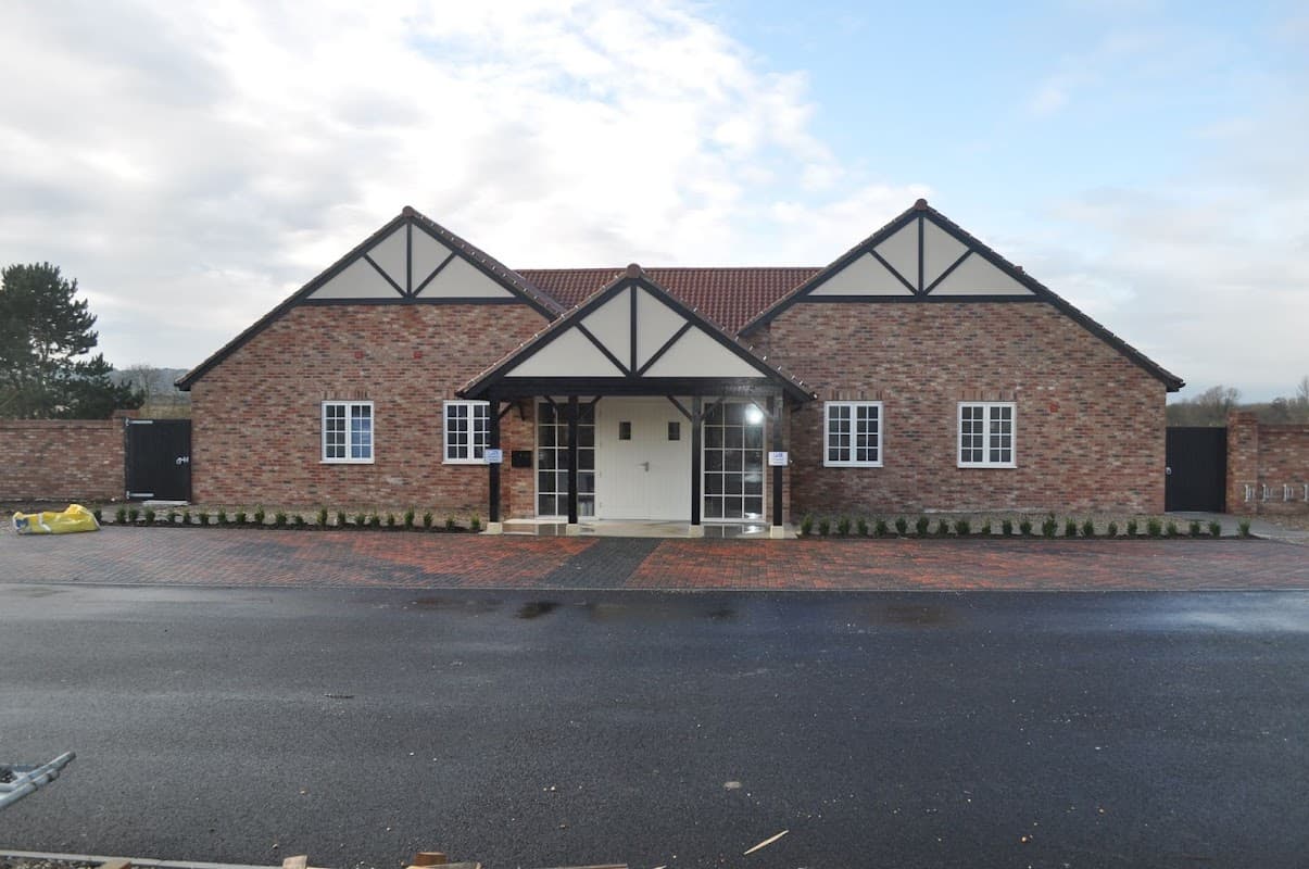 Brick building with a gabled roof, white windows, and a central entrance, surrounded by a paved area and greenery.