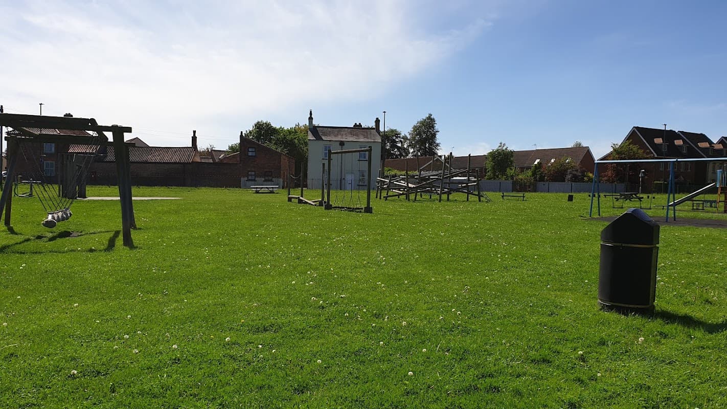 Green park area with swings, climbing structures, and a small building under a clear blue sky.