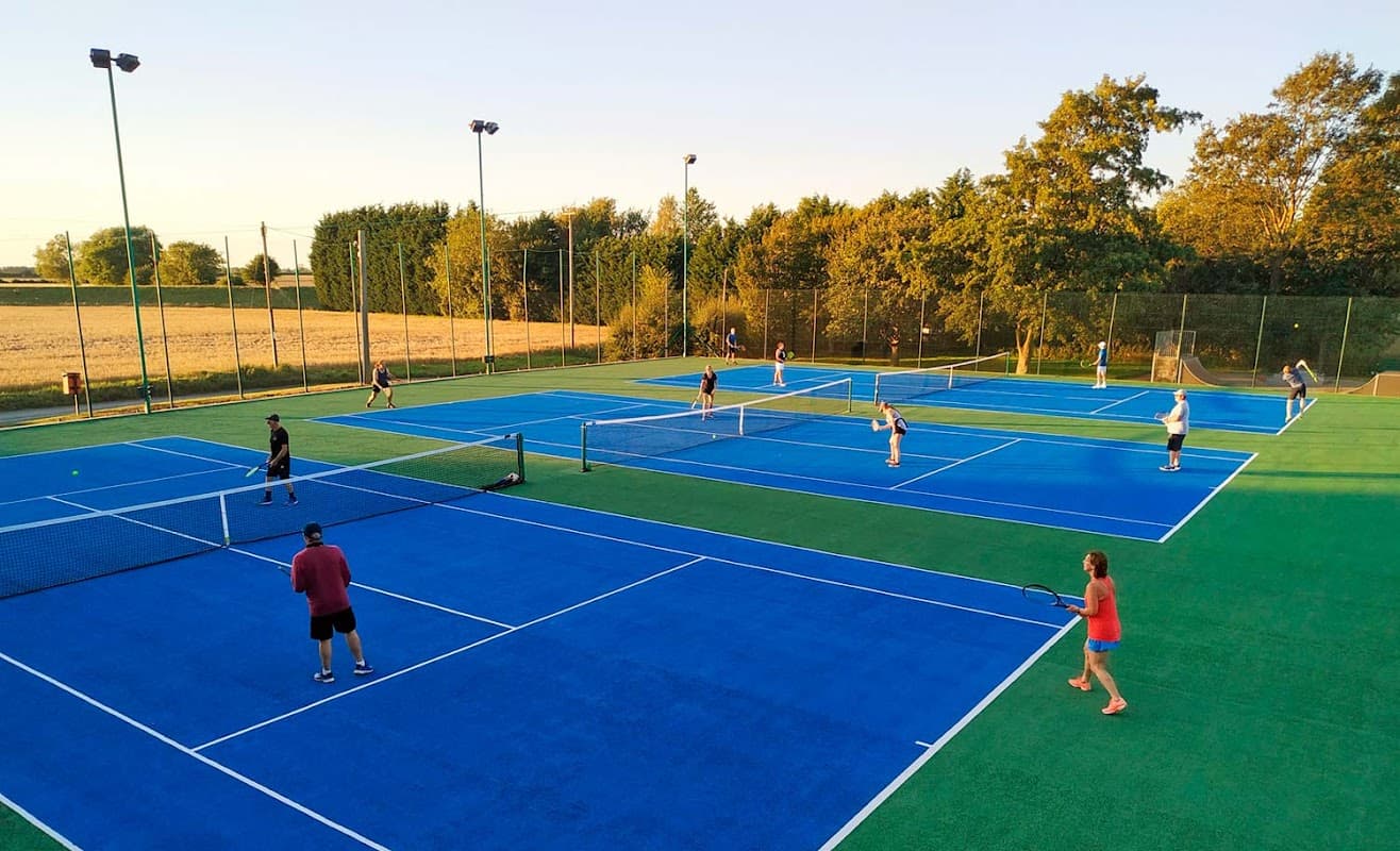Several players engage in tennis on vibrant blue courts surrounded by greenery at Wistow Tennis Club.