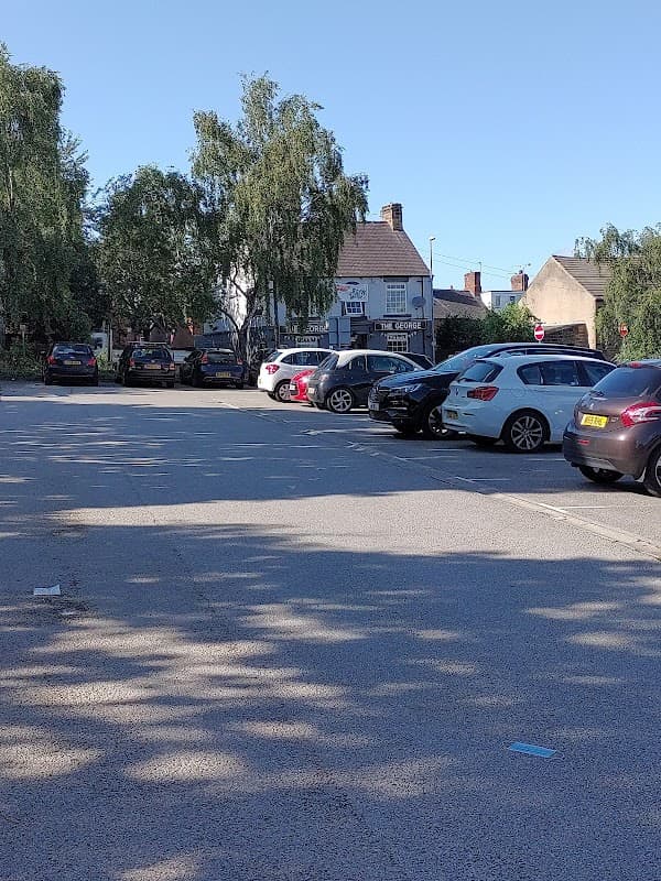 Free parking lot with parked cars, trees, and buildings in Wombwell, Yorkshire under clear blue sky.