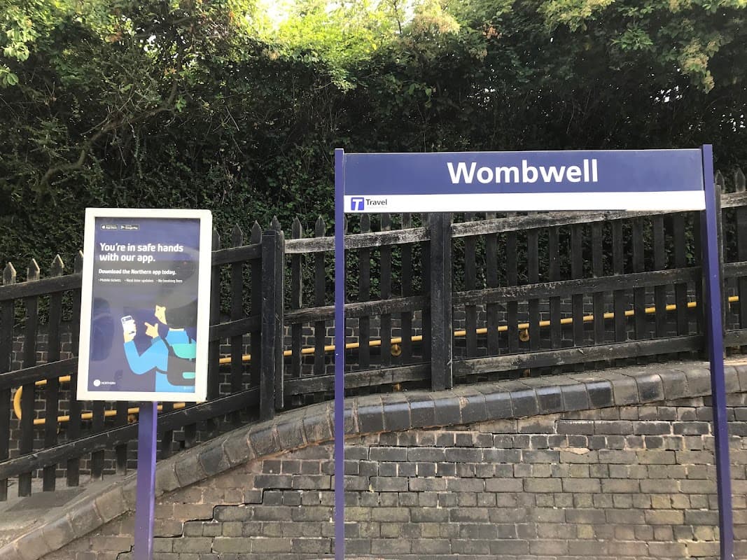 Wombwell bus stop sign with a travel app advertisement nearby, surrounded by greenery and a stone wall.