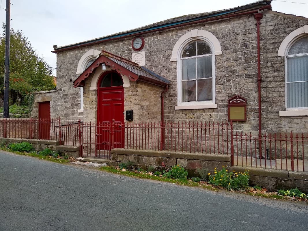 Stone building with red door and fence, clock above entrance, surrounded by greenery in Wrelton, Yorkshire.