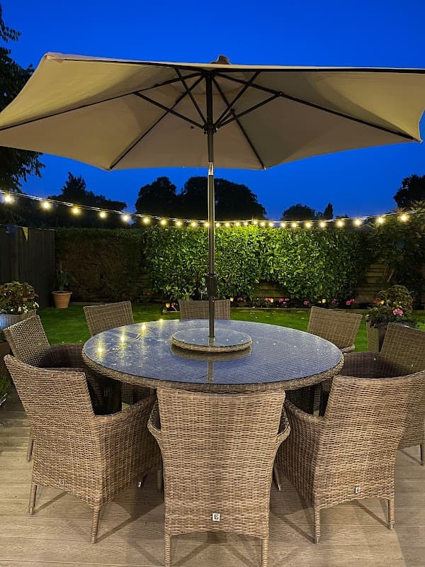 Outdoor dining area with a large round table, wicker chairs, and an umbrella, illuminated by string lights at dusk.
