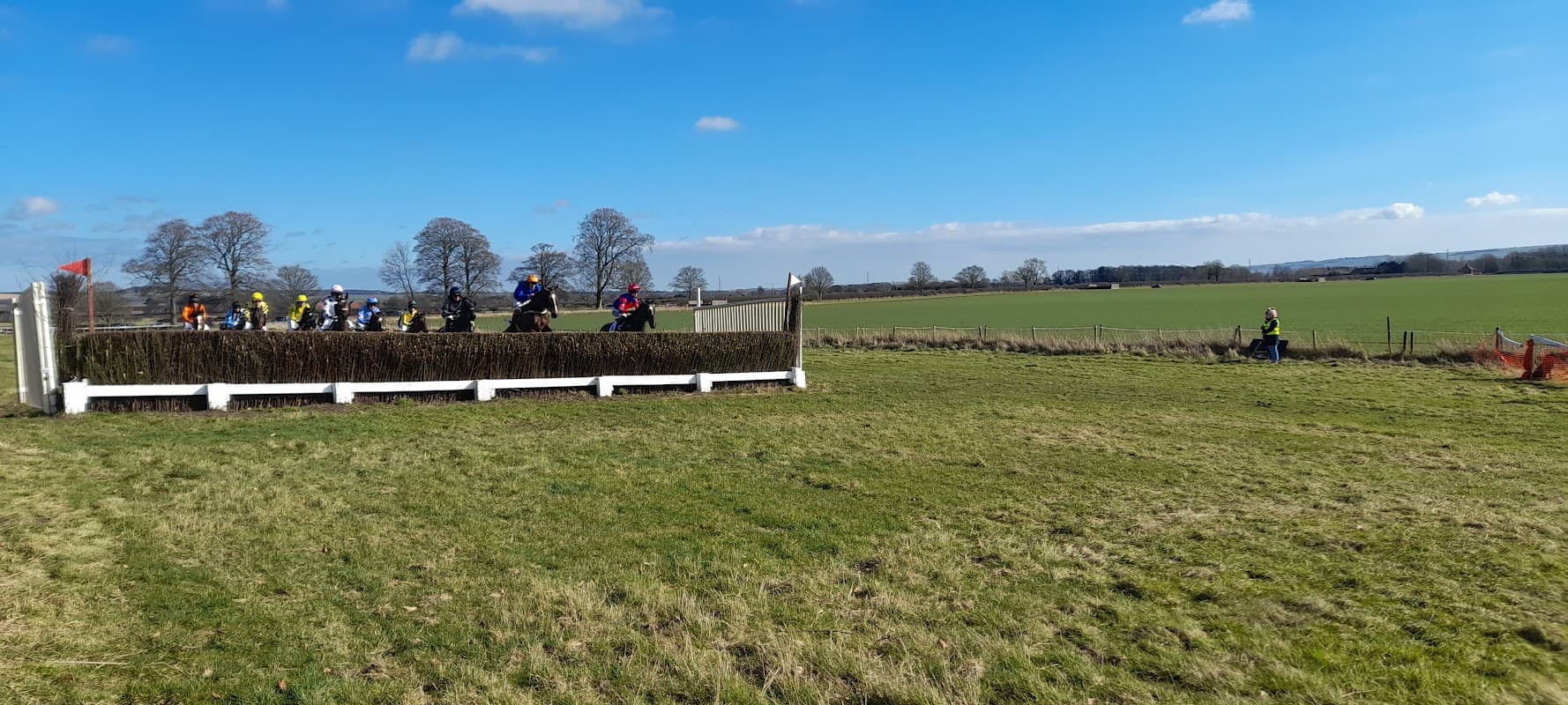 Horses and riders jump a fence in a grassy field under a blue sky, with trees and distant hills in the background.