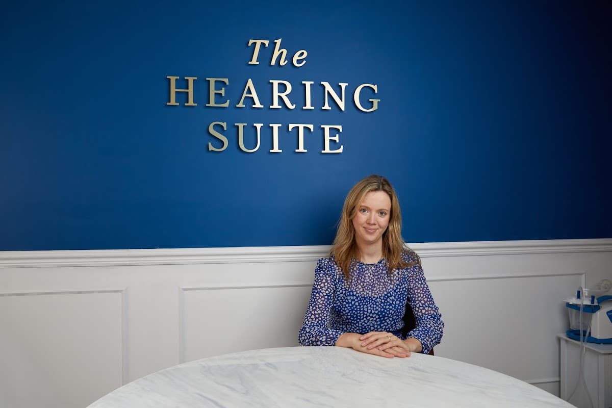 A woman in a blue blouse sits at a marble table in front of a blue wall with "The Hearing Suite" sign.