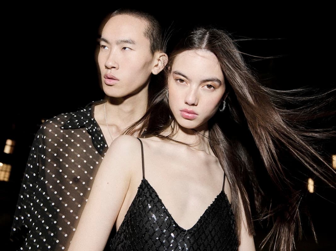 Two models pose closely together, showcasing stylish, modern clothing against a dark background.