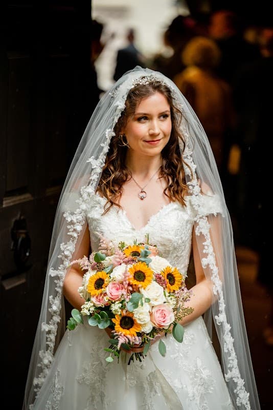 A bride in a lace dress and veil holds a vibrant bouquet of sunflowers and pastel flowers, smiling softly.