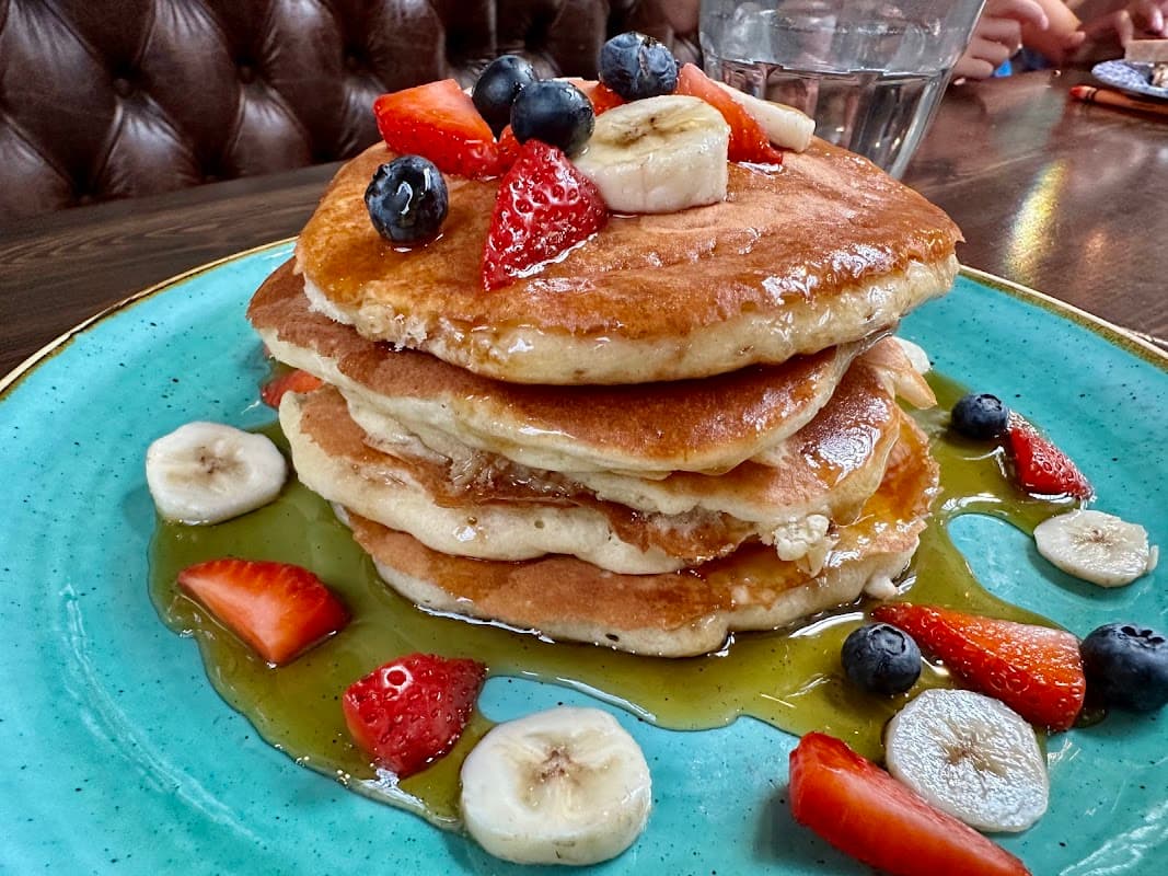 Stack of fluffy pancakes topped with strawberries, blueberries, and banana slices, drizzled with syrup on a teal plate.