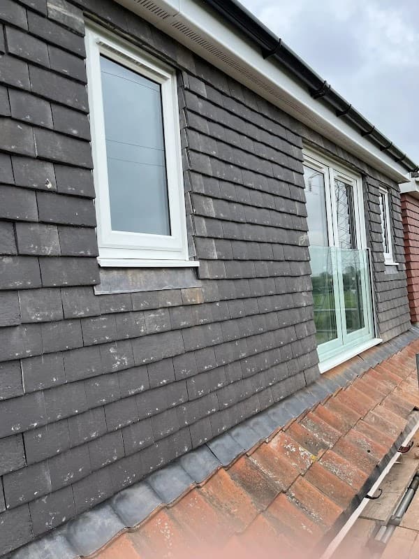 Newly installed windows on a grey shingled wall, with a roof edge visible below and cloudy sky above.