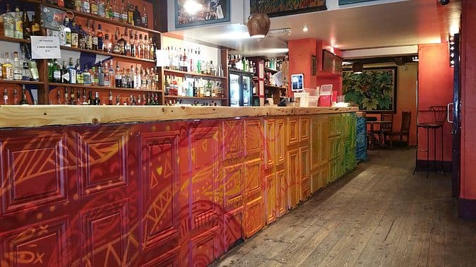 Colorful bar with a vibrant mural, shelves stocked with various spirits, and wooden flooring in a lively setting.
