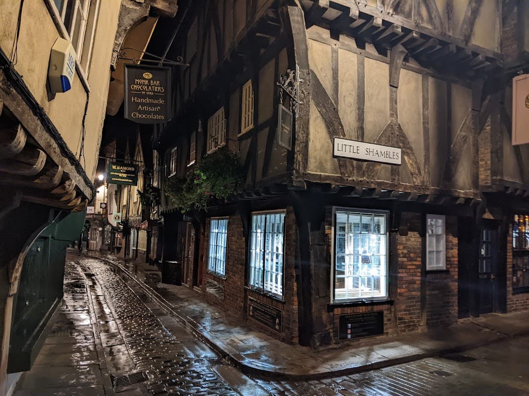 Historic narrow alley with timber-framed buildings, signage for "Little Shambles," and shop windows reflecting light.