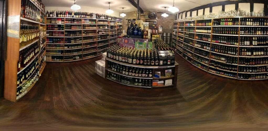 A cozy off-licence with wooden floors, shelves filled with various bottles of beer and spirits, and warm lighting.