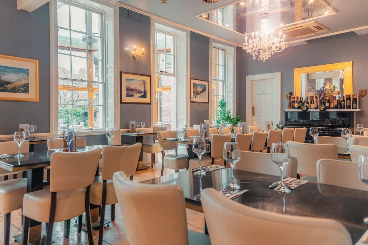 Elegant dining area with white chairs, tables set with glassware, large windows, and a chandelier.