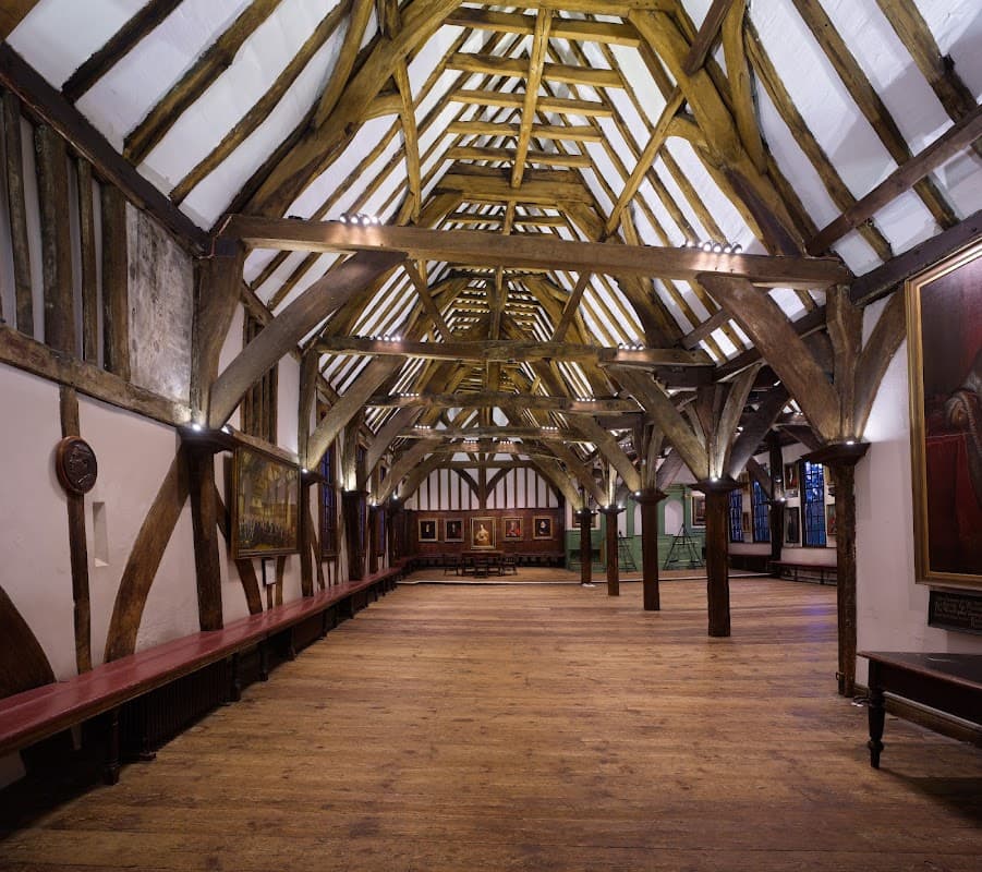 Spacious hall with wooden beams, benches along the walls, and framed paintings in a historic setting.