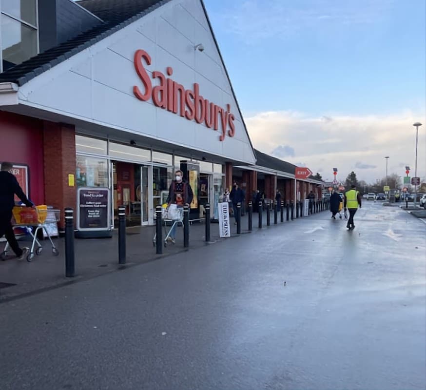 Sainsbury's storefront with shoppers and trolleys outside, cloudy sky, and parking area visible.