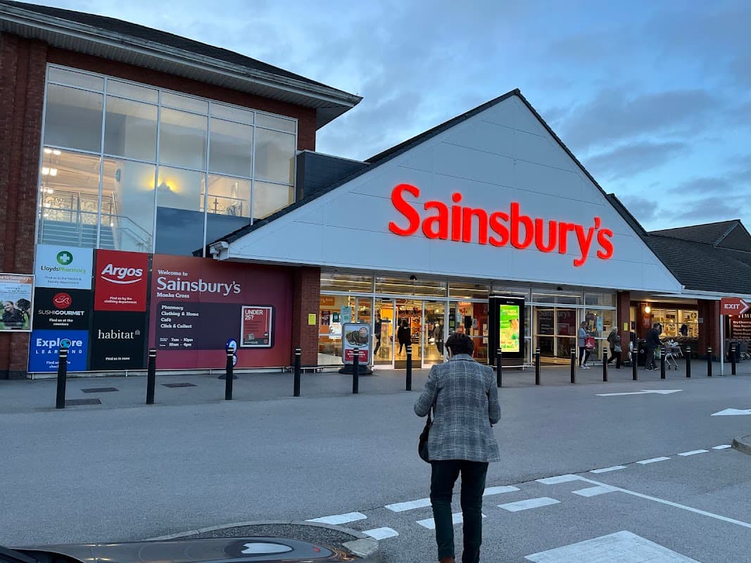 Sainsbury's storefront at Monks Cross Shopping Park, with various store signs and a shopper approaching.
