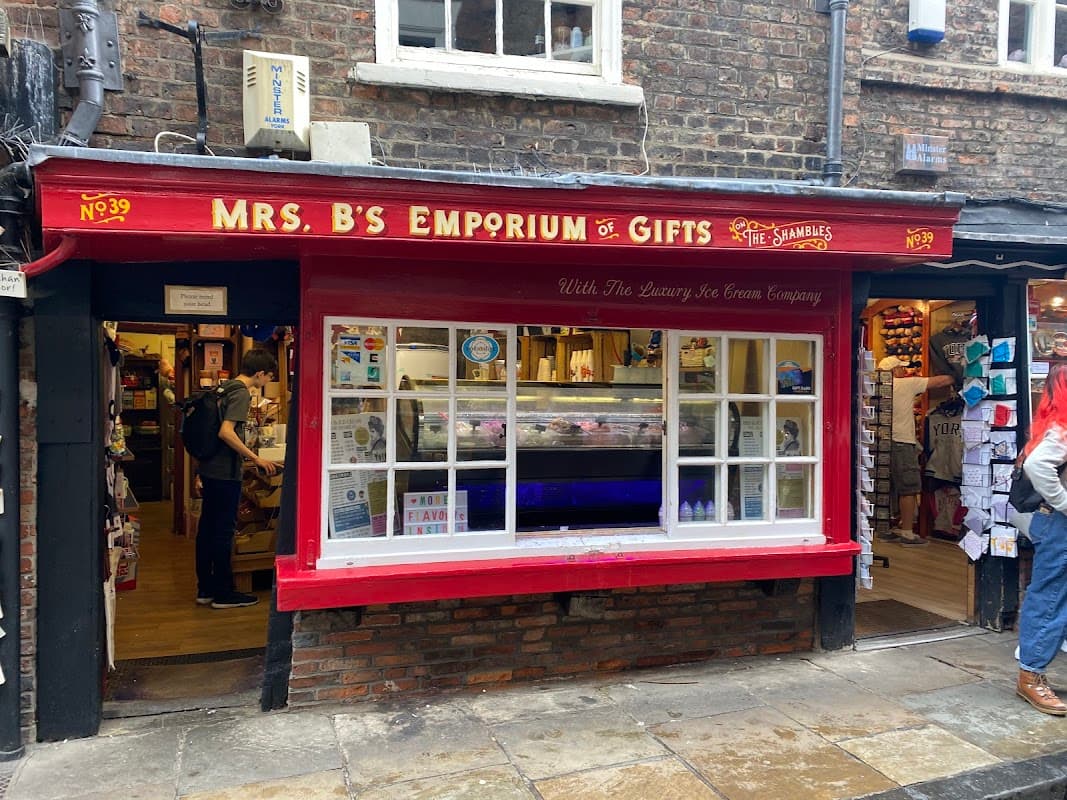 Red storefront of Mrs B's Emporium of Gifts with large window displaying gifts and ice cream, cobbled street outside.