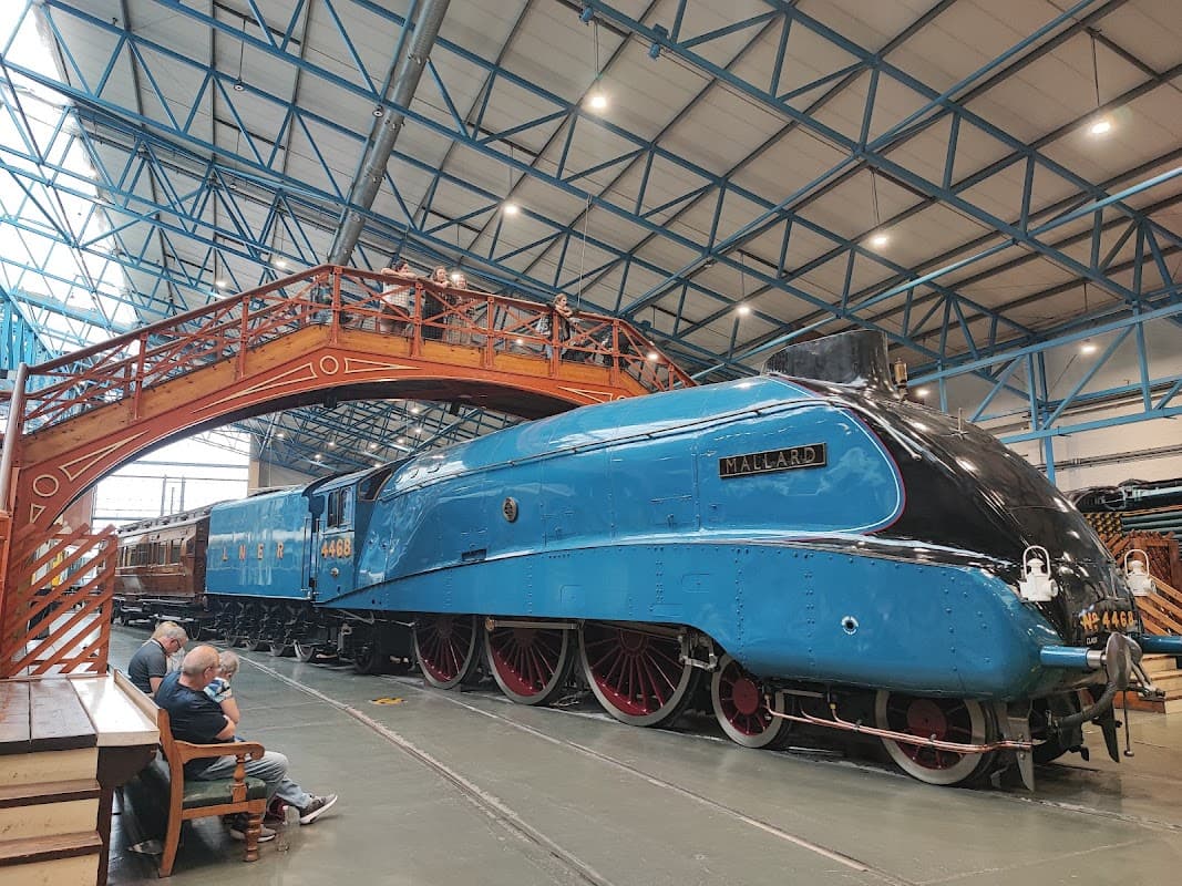 Blue steam locomotive "Mallard" displayed indoors beneath a high ceiling, with wooden bridge and visitors nearby.