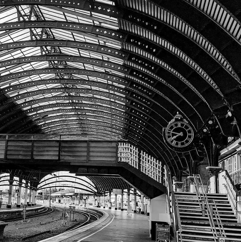 Historic train station interior with arched metal roof, clock, and stairway leading to platforms. Black and white photo.