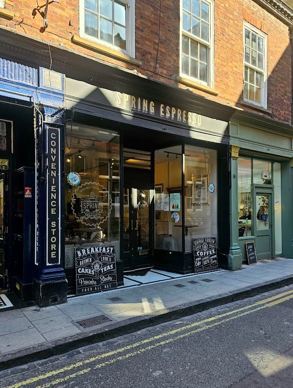 Charming café entrance with a sign for "Spring Espresso," surrounded by brick buildings and a convenience store nearby.