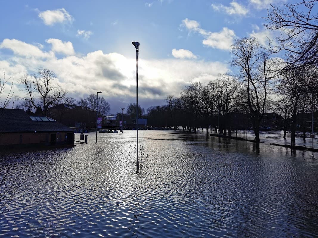 Flooded car park with standing water, trees in the background, and a cloudy sky with sunlight breaking through.