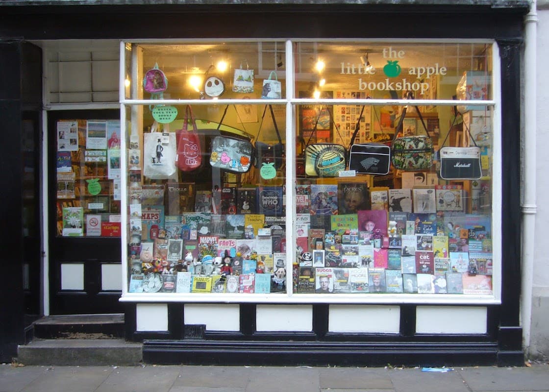 Colorful window display of "The Little Apple Bookshop" featuring various books, bags, and decorative items.