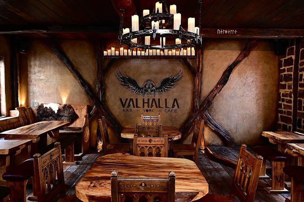 Rustic bar interior with wooden tables, ornate chairs, and a large chandelier above. Valhalla logo on the wall.