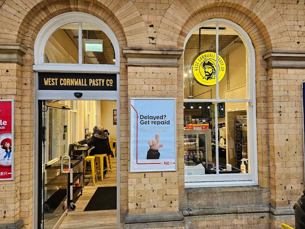 West Cornwall Pasty shop entrance with large windows, yellow signage, and a poster about delays. Customers inside.
