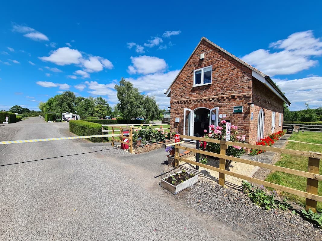 Brick building with a gabled roof, surrounded by greenery, flowers, and a gravel path, under a blue sky with clouds.