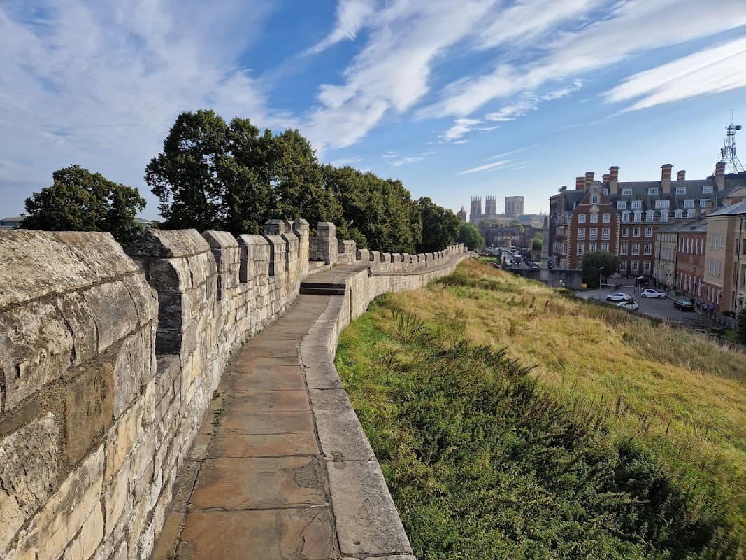 Stone walls of York City Walls with a pathway, lush greenery, and historic buildings under a blue sky.