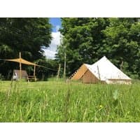 Canvas bell tent with triangular entrance, pitched on grassy field, surrounded by trees, with a wooden picnic table nearby.