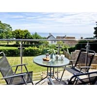Glass balcony table set with coffee pot and cups, overlooking lush garden and distant sea view, surrounded by metal chairs.