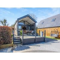 Modern grey cabin with glass doors, elevated porch, and sloped roof, surrounded by greenery and adjacent wooden structure.