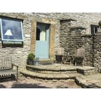 Stone cottage with blue door, wooden chairs, and table on a stone patio, surrounded by rustic stone walls.