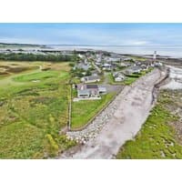 Coastal holiday cottage "Driftwood" with white exterior, near rocky shoreline and green landscape, by cottages.com.