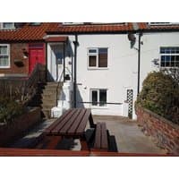 White cottage with red roof, red door, and outdoor wooden picnic table on stone patio.
