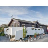 Brown wooden cottage with white fenced patio, potted plants, and gravel path under a blue sky.