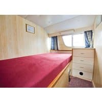 Houseboat bedroom with wood paneling, red bedspread, small window, blue curtains, and a dresser with four drawers.