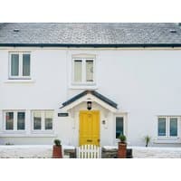 White cottage with a slate roof, bright yellow door, and small front garden with potted plants.