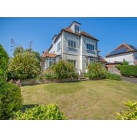 White three-story beach house with red roof, large windows, surrounded by lush garden and clear blue sky.