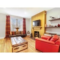 Living room with red sofa, plaid curtains, wooden floor, rustic chest coffee table, wall-mounted TV, and decorative shelves.